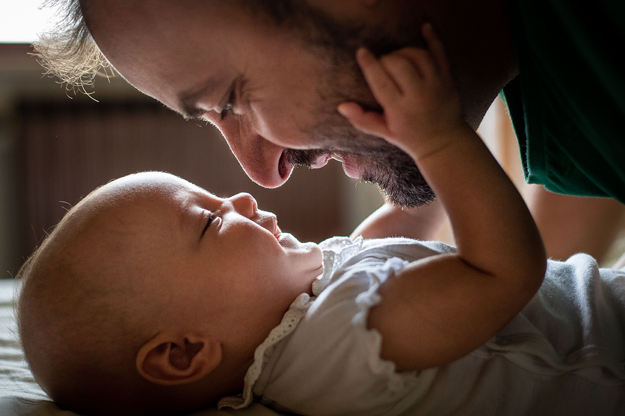 Ein Vater beugt sich lächelnd über sein Baby, das auf dem Rücken liegt und nach seinem Gesicht greift.