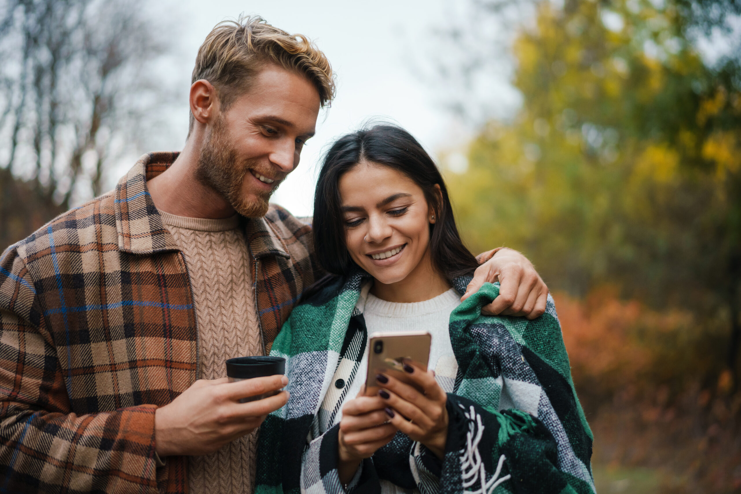 Junges Paar schaut gemeinsam lächelnd auf ein Smartphone, herbstliche Umgebung im Hintergrund.