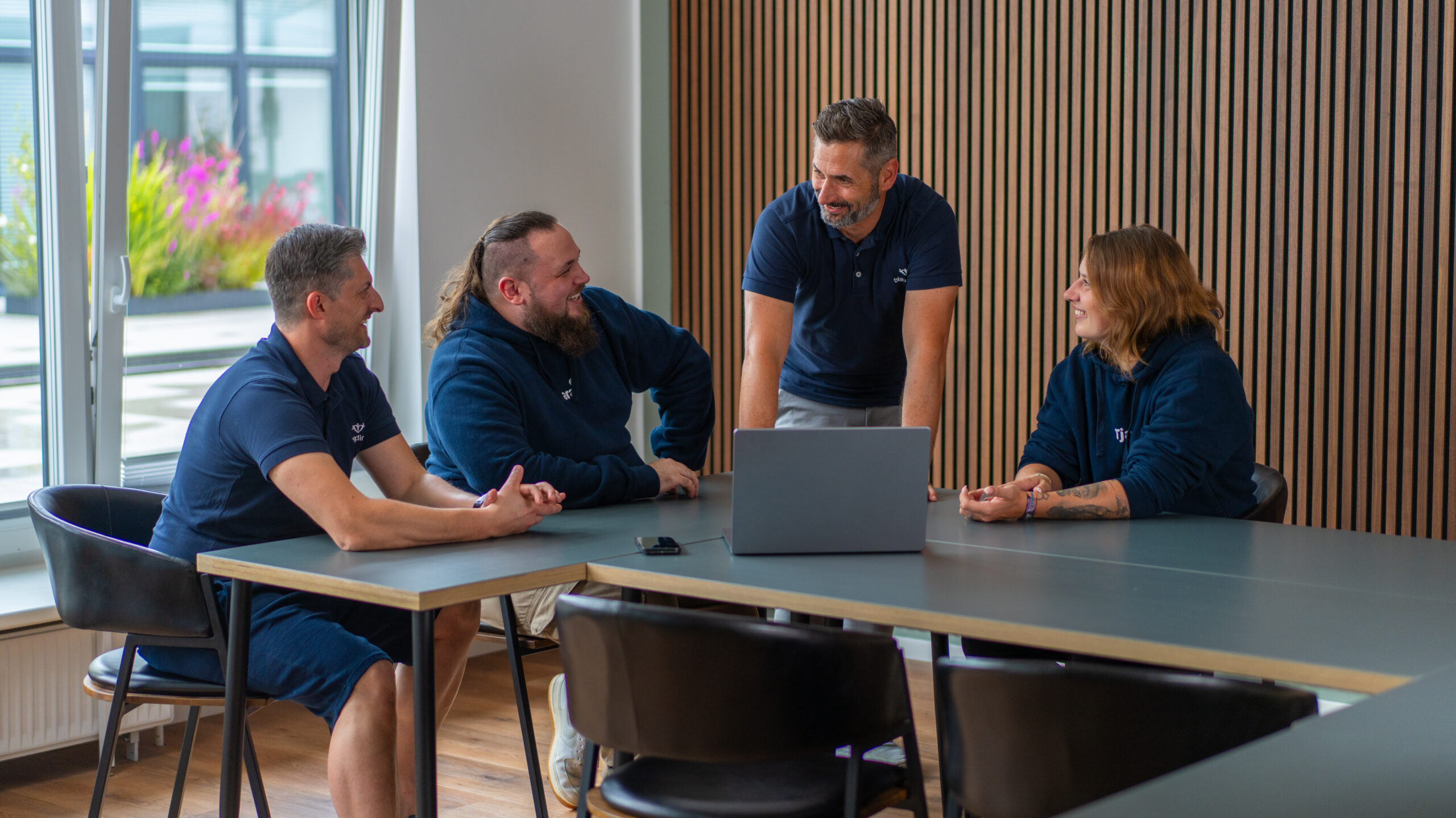Vier Mitarbeitende in dunkelblauer Kleidung besprechen sich locker am Konferenztisch mit Laptop