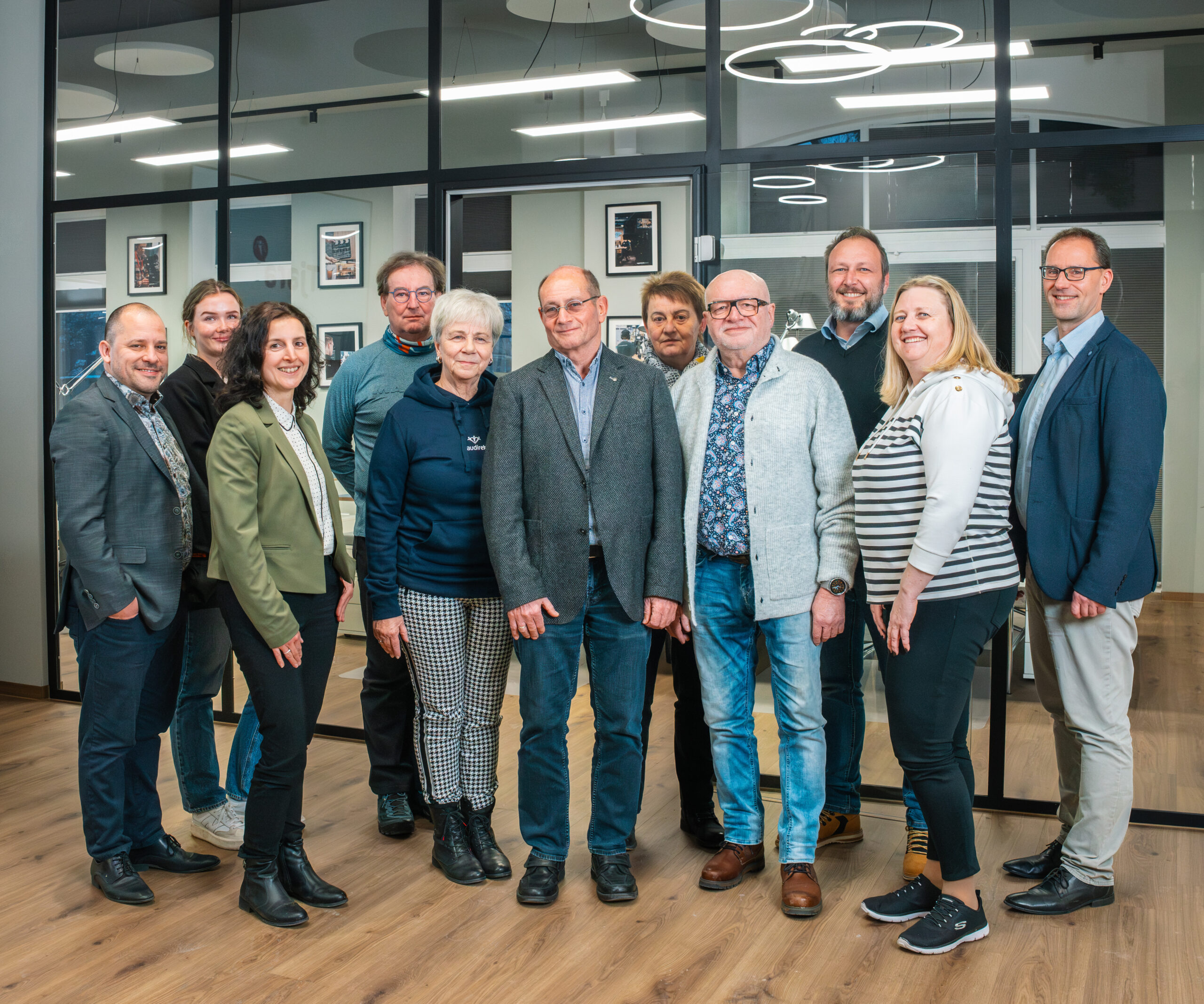 Elfköpfiges Team aus Frauen und Männern steht lächelnd nebeneinander in einem modernen Büro mit Glaswänden und runden Lichtelementen